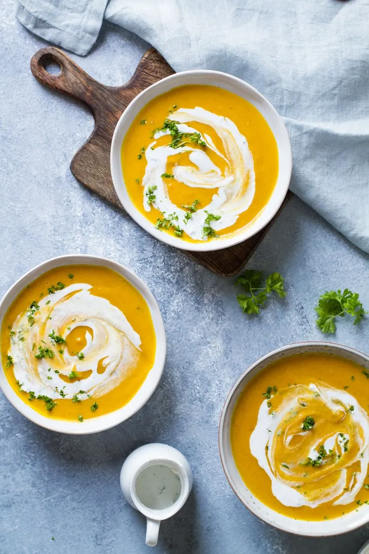 Three bowls with curry coconut carrot soup with ginger cream and parsley. Blue background.
