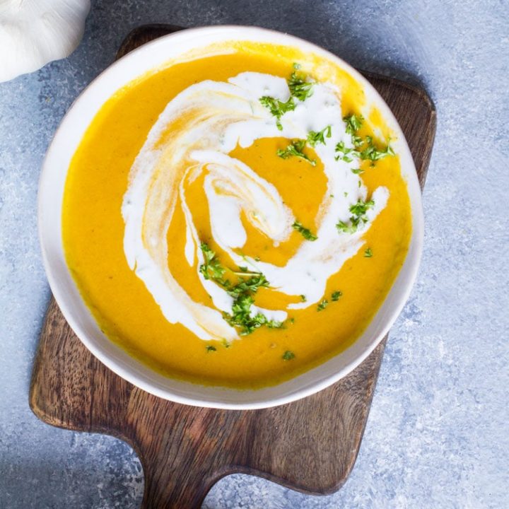 White bowl with curry coconut carrot soup on a wooden cutting board on a blue table.