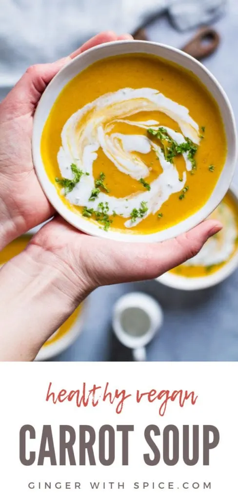 Hands holding a bowl with curry coconut carrot soup garnished with ginger cream and parsley. Pinterest pin.