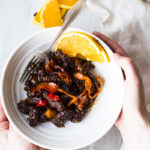 Hands holding a bowl with orange Szechuan beef and carrots.