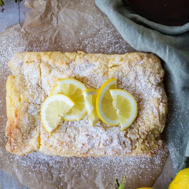 Lemon Cake with lemon slices and powdered sugar on top. Background is parchment paper and a green towel,