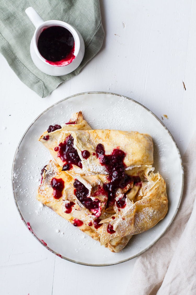 Flat lay: A stack of crepes filled with lemon cream cheese and blackberry jam. White plate, white background and white mug with blackberry jam.