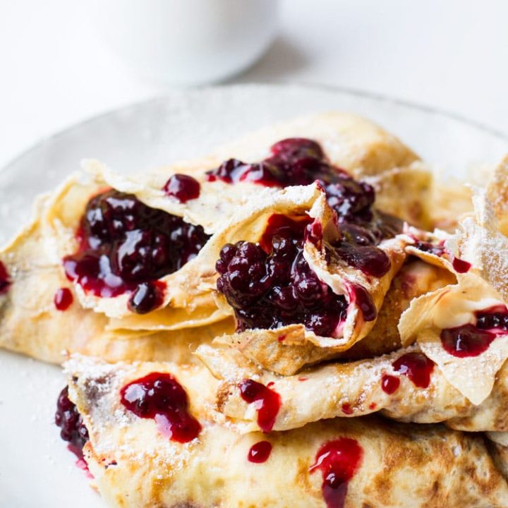 A stack of crepes filled with lemon cream cheese and blackberry jam. White plate, white background and white mug with blackberry jam.