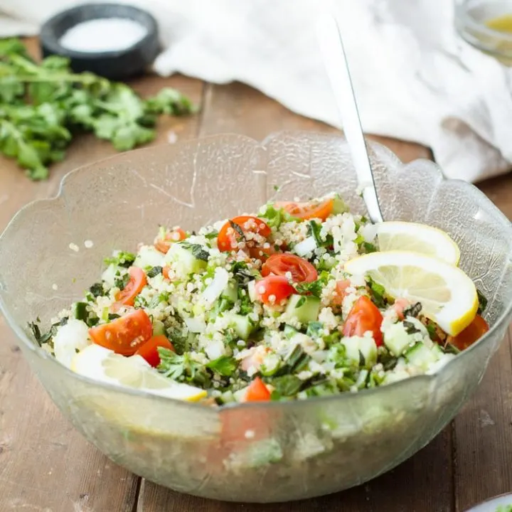 Tabbouleh and lemon wedges in a large glass bowl. Ingredients and towel blurred in the background.