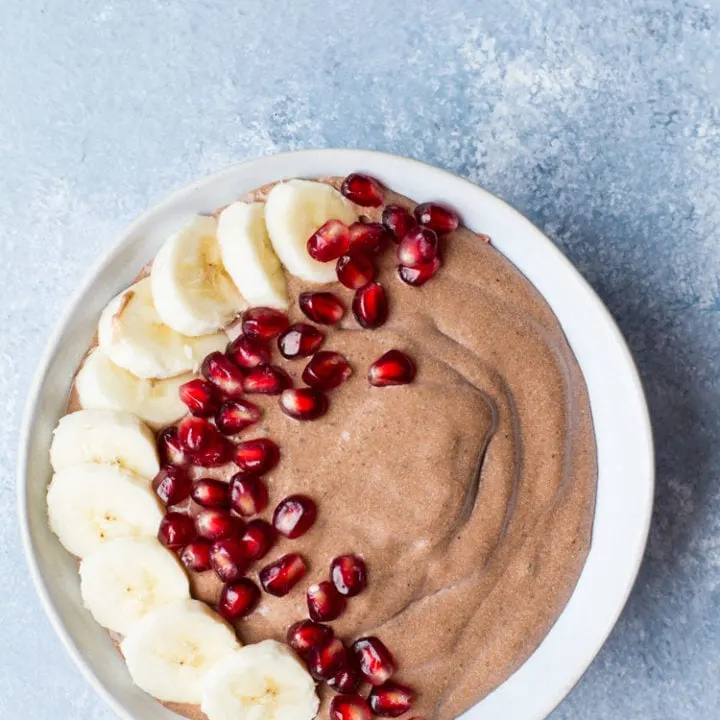 Bowl with chocolate chia pudding, decorated with banana slices and pomegranate to the left. Blue background, flatlay.