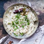 Bird's eye view of a metal pan with Indian rice with cilantro, cinnamon stick and bay leaf. Rusty background and nude colored cloth.