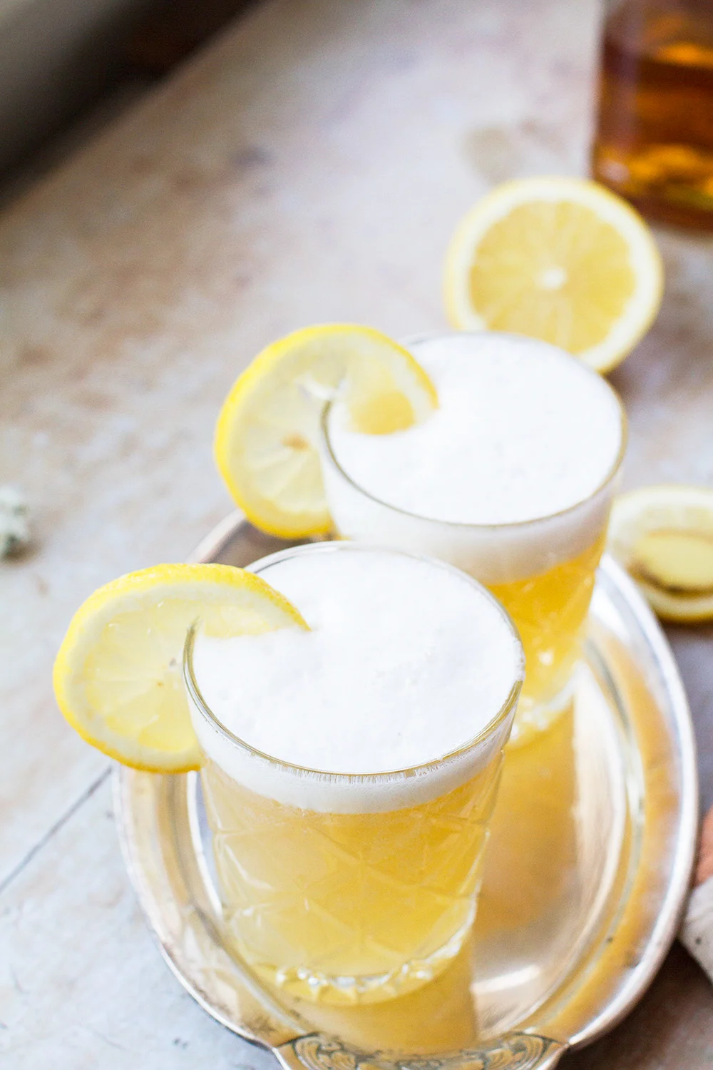 Two glasses with whiskey sour and a frothy top, garnished with lemon slices.