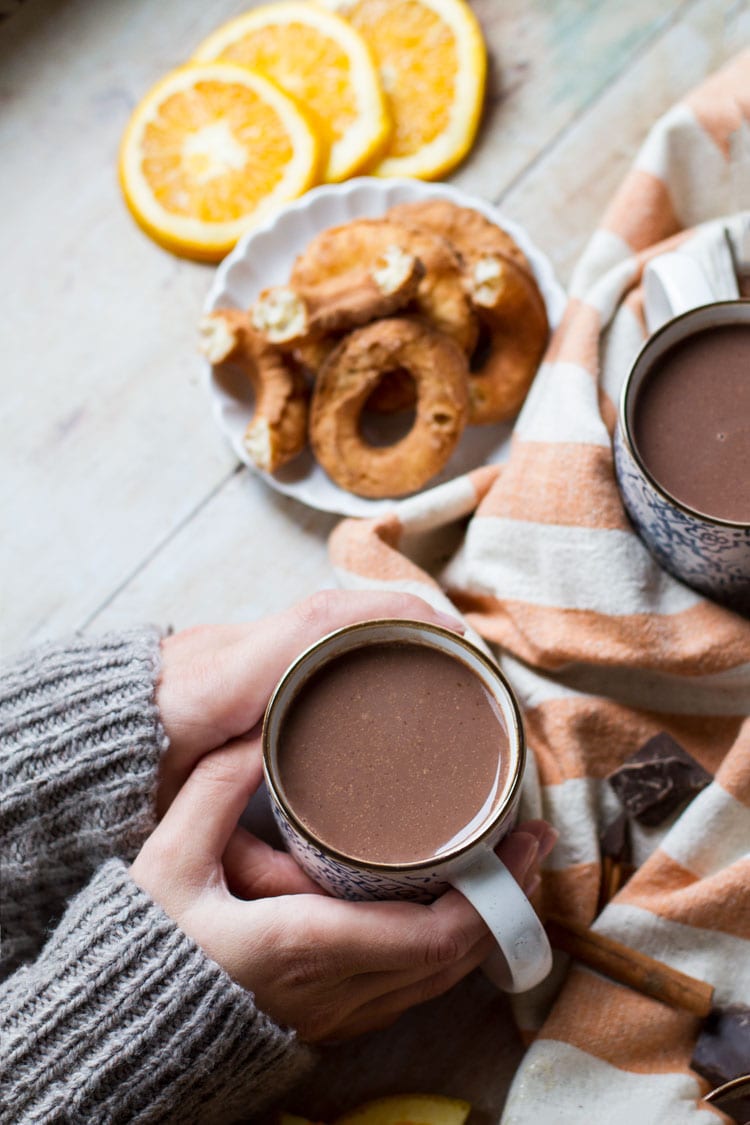Hands holding a cup of orange hot chocolate. Donuts in the background.