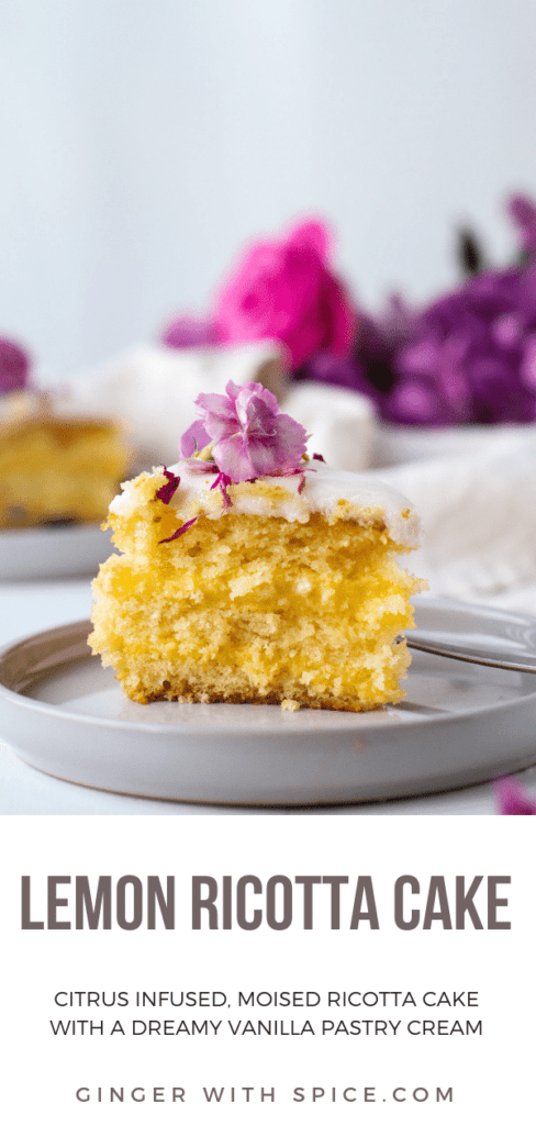 Yellow slice of cake on a grey plate with a purple flower on top. More purple flowers in the background.