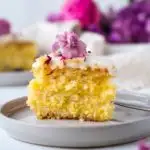 Yellow slice of cake on a grey plate with a purple flower on top. More purple flowers in the background.