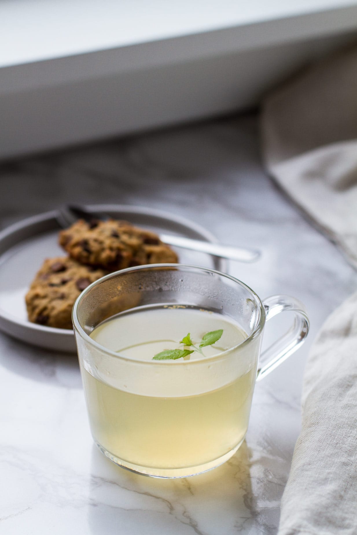 Clear mug with pale yellow drink and lemon verbena garnish. Cookies in the background.