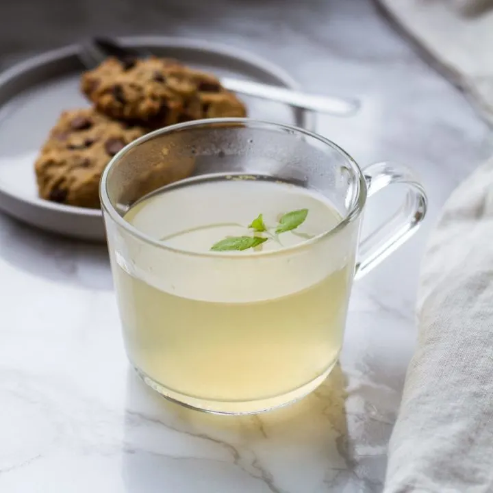 Clear mug with pale yellow drink and lemon verbena garnish. Cookies in the background.