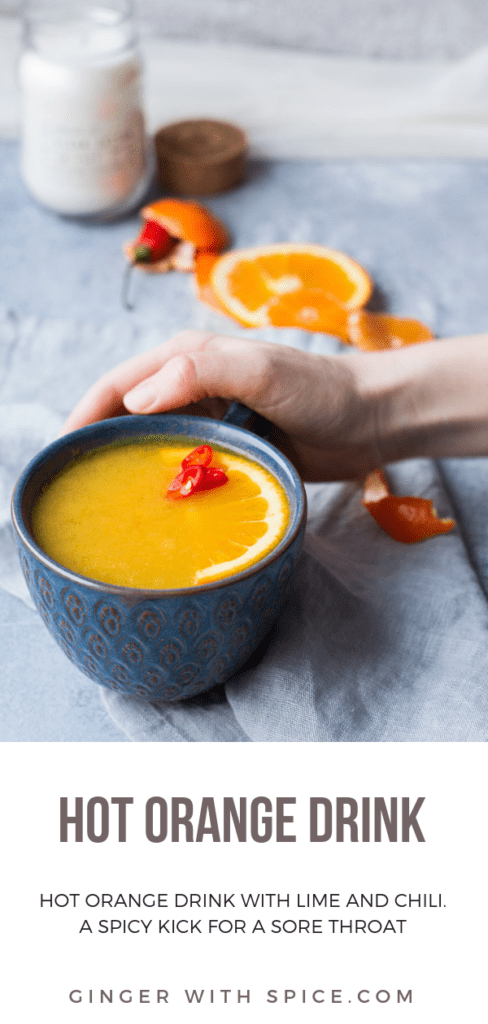 Blue cup with hot orange drink, half an orange slice and three chili slices, hand holding cup. Blue background and oranges and orange peel blurred out in the background.