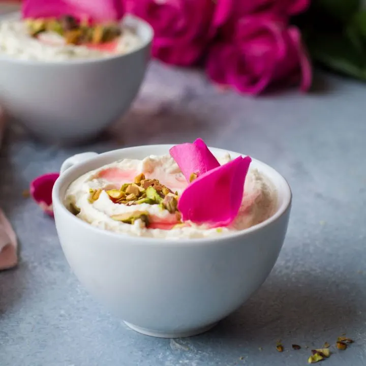 White cup with white mouse, chopped pistachios and rose petals. Roses and another cup blurred in the background. Blue table.