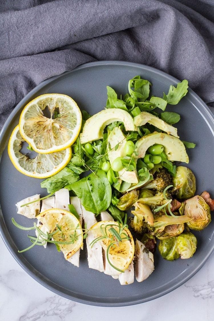 Flat lay of baked lemon chicken with green spring salad and lemon slices, dark grey plate. Turned to be horizontal.