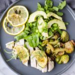 Flat lay of baked lemon chicken with green spring salad and lemon slices, dark grey plate. Square crop.