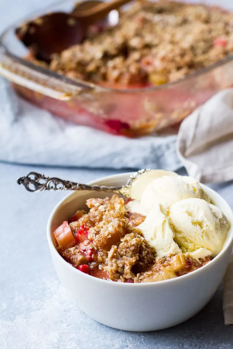 White bowl with rhubarb crisp and two scoops of vanilla ice cream. The complete dish in the background.