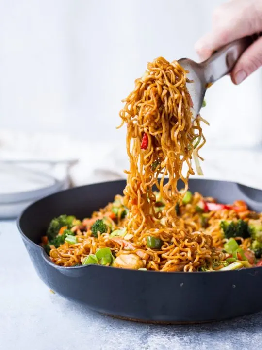 A metal tong holding up noodles from vegetarian lo mein in skillet.