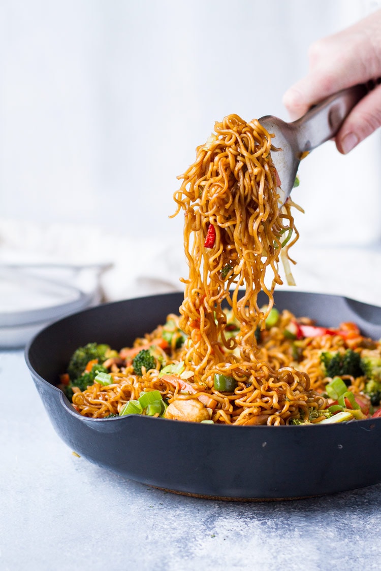 A metal tong holding up noodles from vegetarian lo mein in skillet.