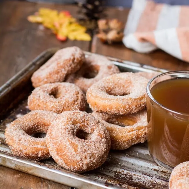 Donuts coated in cinnamon sugar and apple cider in a clear cup on a metal pan.