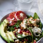 Big bowl of lemon chicken winter salad, topped with sliced avocado and blood orange. Glass of water in the background.
