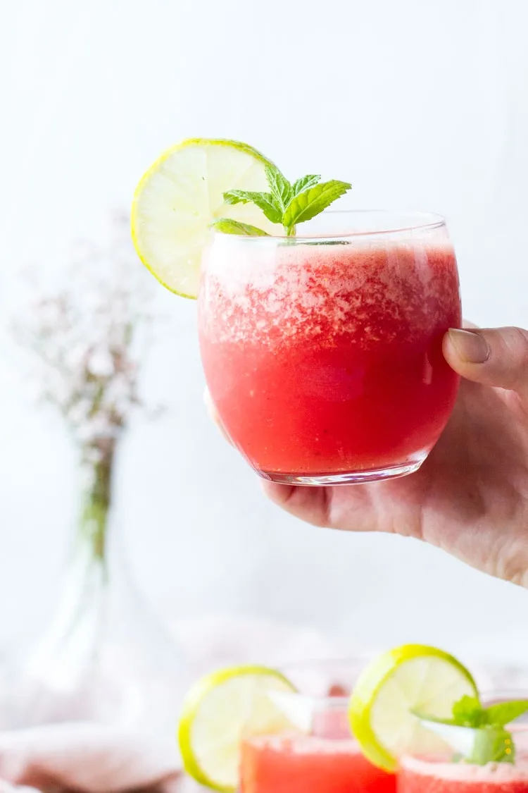 Hand holding a glass of watermelon limeade, garnised with lime and mint.