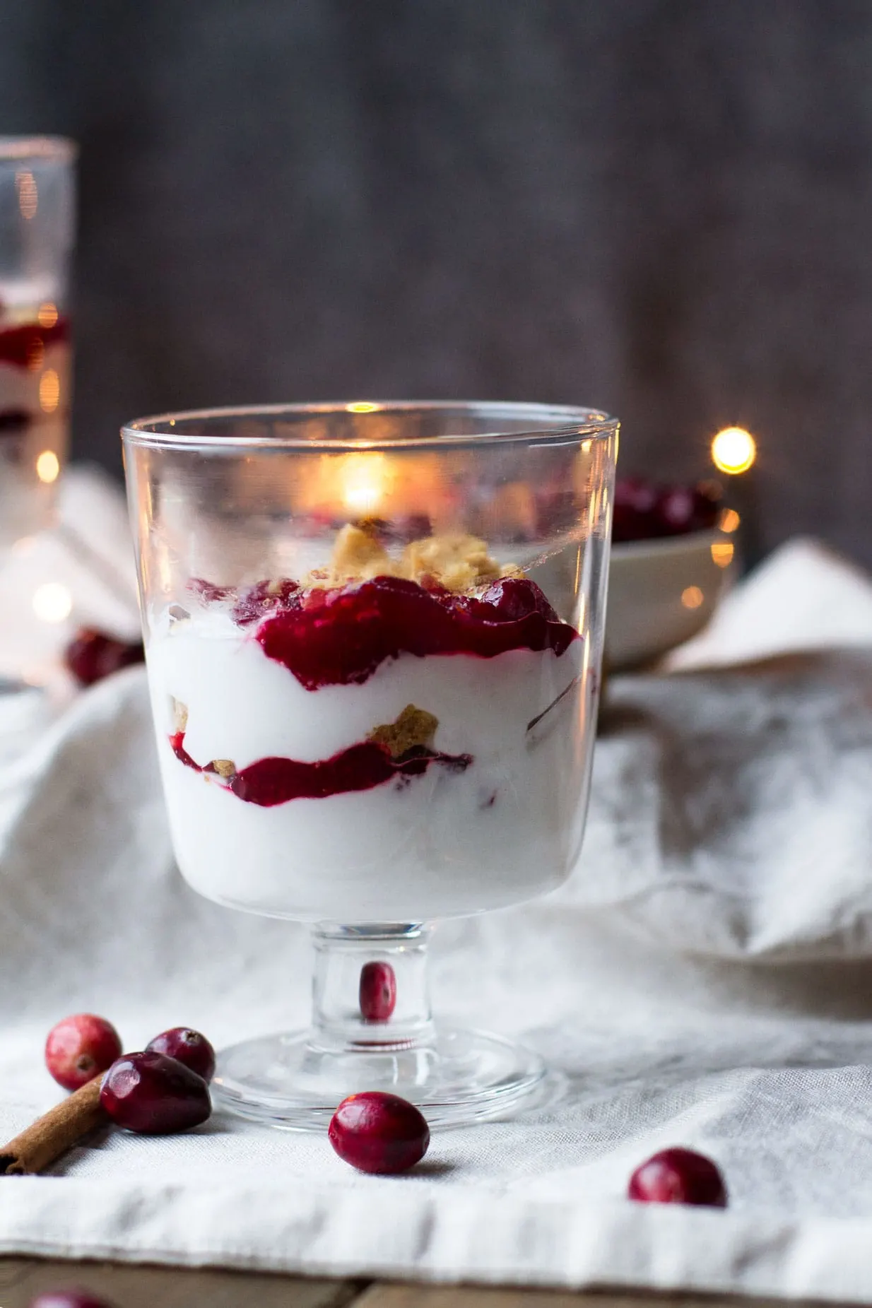 Glass with layers of cranberry sauce, whipped coconut cream and ginger nut cookies. Lights in the background.
