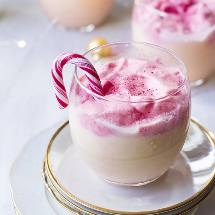 Round glass with eggnog and fluffy pink dalgona, garnished with candy cane.