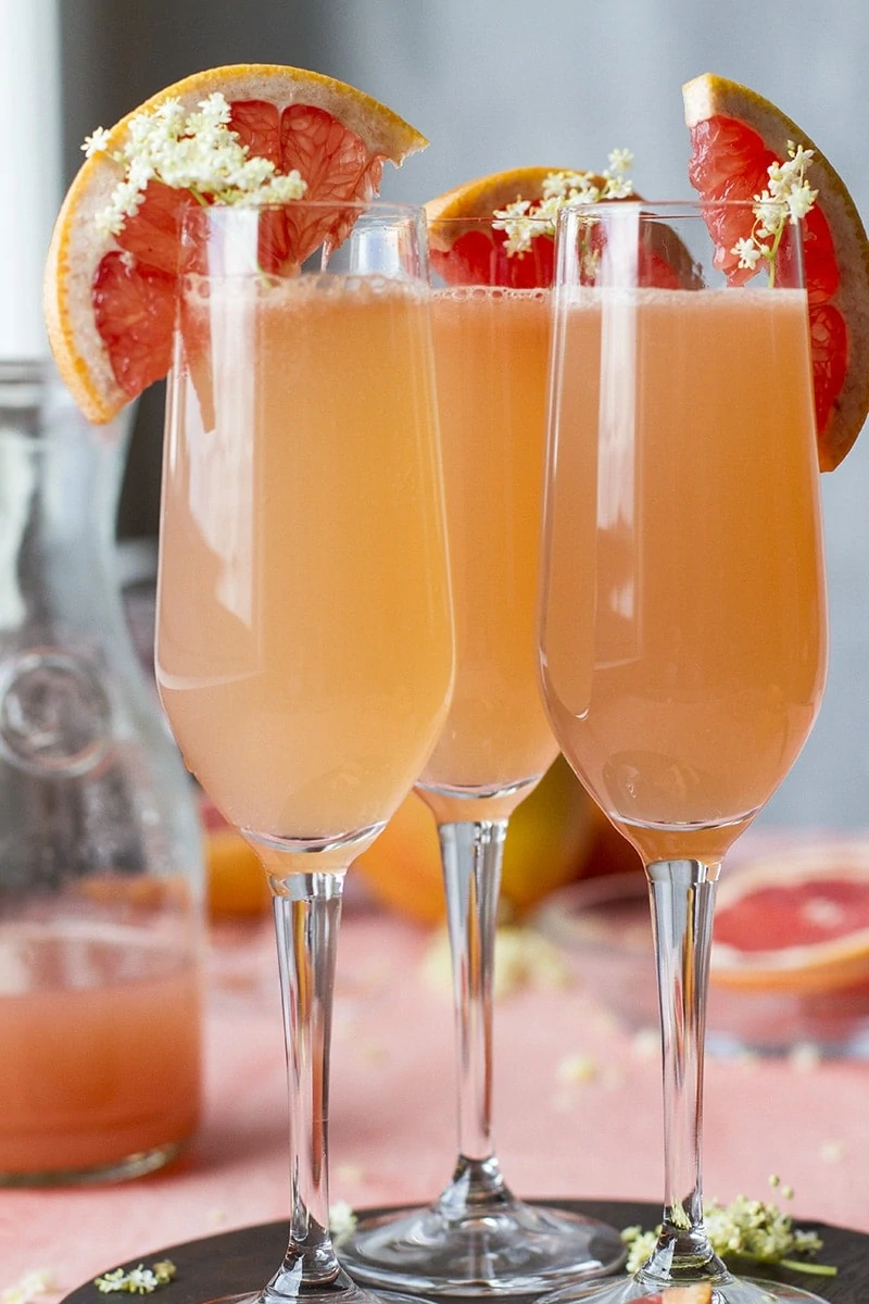 Close-up of three champagne flutes with grapefruit mimosa. Garnished with sliced grapefruit and elderflower.