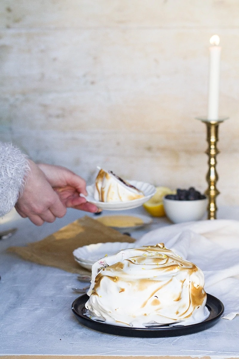 The whole lemon blueberry cake in the front and hands holding a plate with a slice in the background.