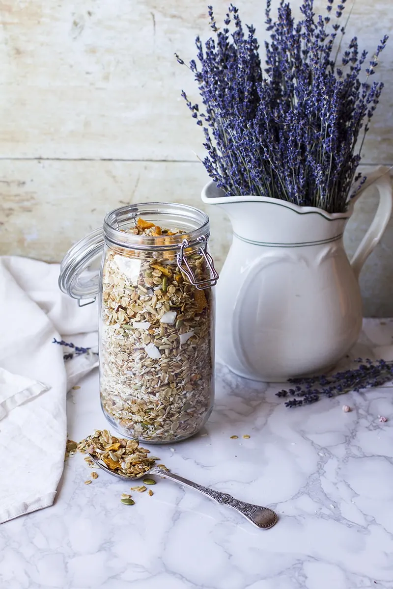 A big glass jar with granola and a vase with lavender flowers in the background.