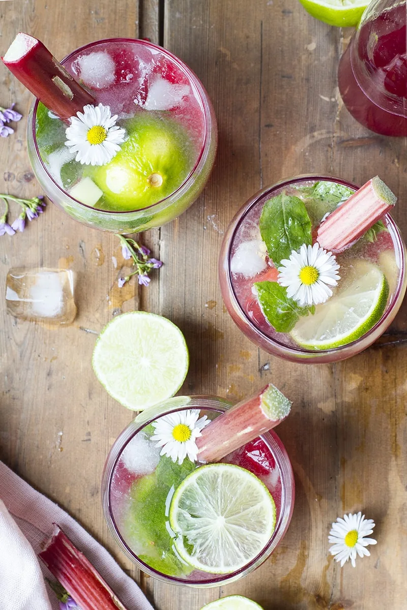 Three red cocktails seen from above, garnished with lime slices, rhubarb stalks and small flowers.