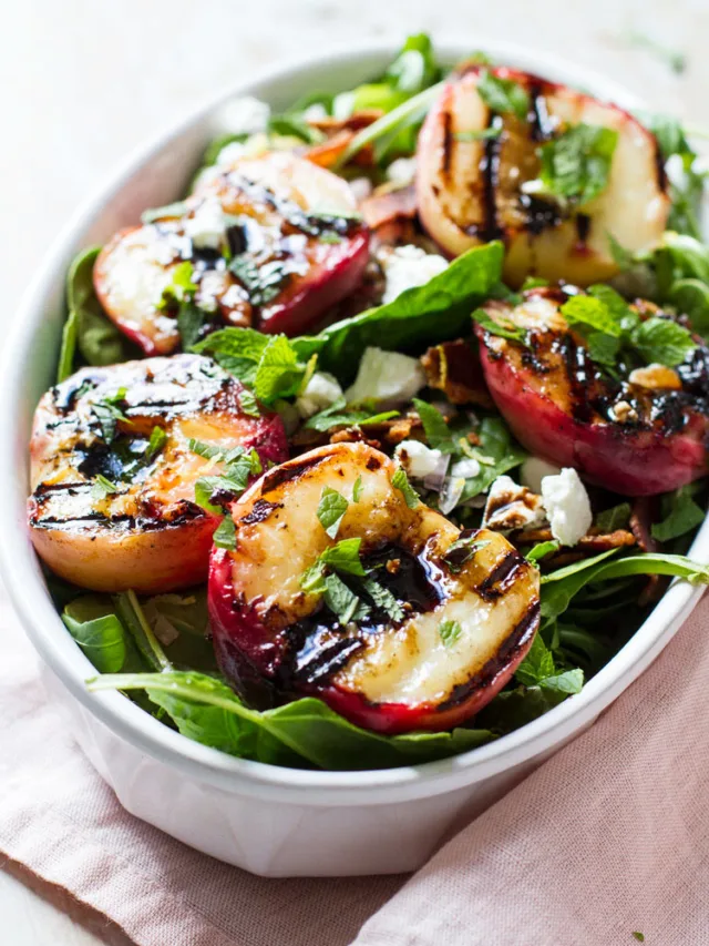 Grilled peaches on top o a salad in a white bowl.
