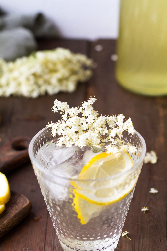 A glass with elderflower cordial, garnished with fresh elderflowers and a lemon wedge.