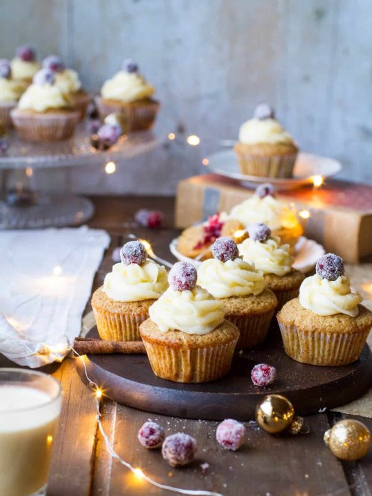 Gingerbread cupcakes on a wooden plate, topped with eggnog icing and sugar cranberries. Sparkling lights.
