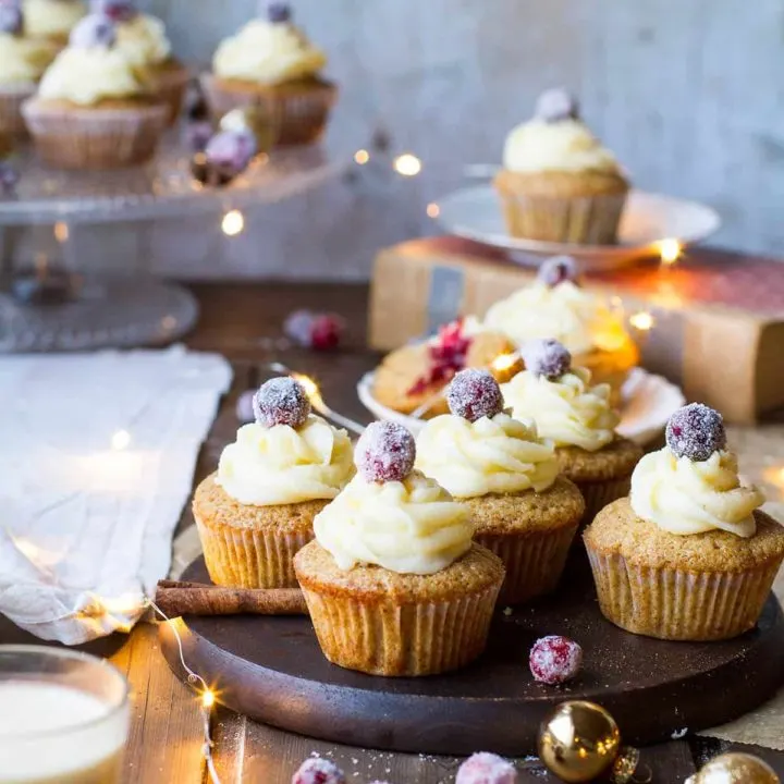 Gingerbread cupcakes on a wooden plate, topped with eggnog icing and sugar cranberries. Sparkling lights.