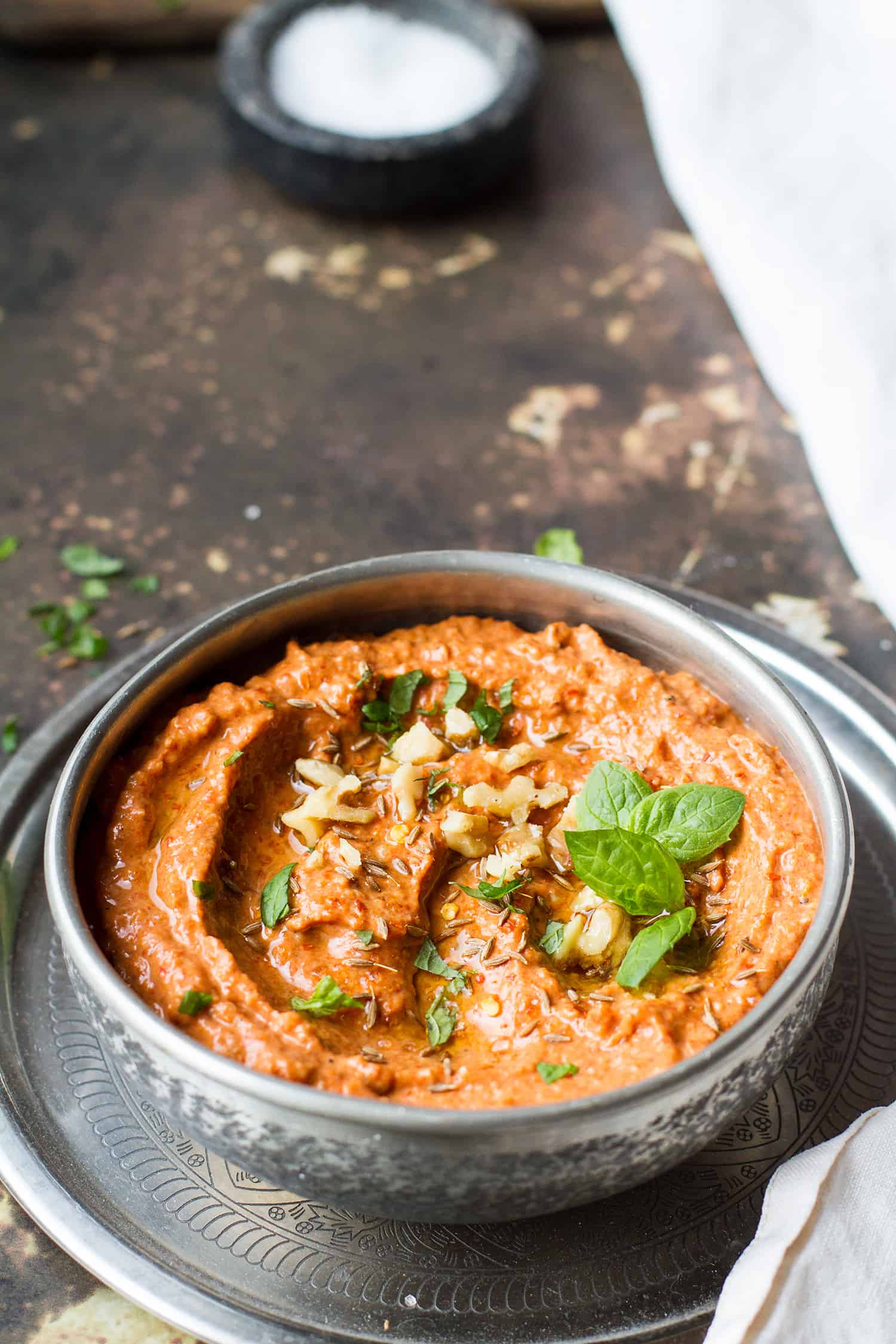 Muhammara dip in a metal bowl, with nuts and herbs as toppings.