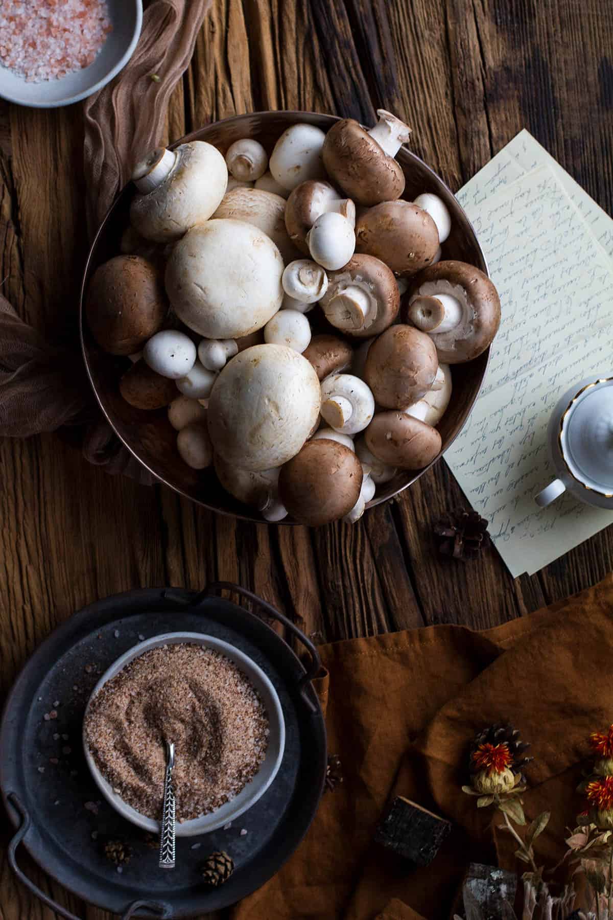 A big bowl of various mushrooms and a small bowl with mushroom salt on a large wooden table.