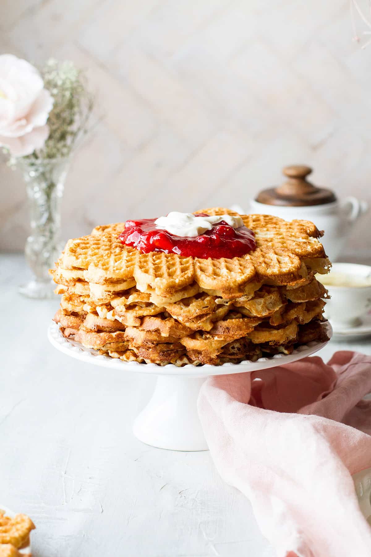 A tall stack of waffles on a cake stand, topped with strawberry jam and sour cream.