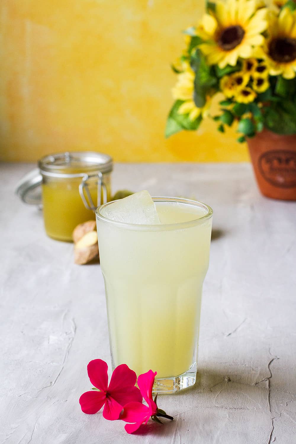 Homemade ginger beer in a large glass, pink flower in the foreground and yellow background.