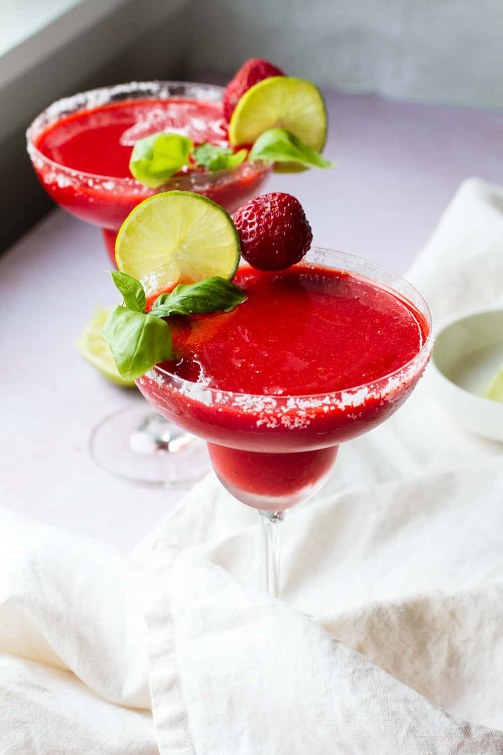 Frozen strawberry margarita in a margarita glass, garnished with lime wheel, strawberry and fresh basil.