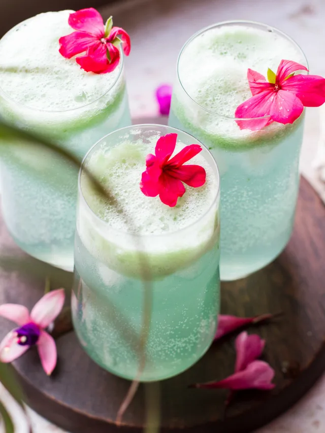 Three tall glasses with turquoise cucumber gin and tonic and fresh pink flowers, some palm leaves blurred out in front.