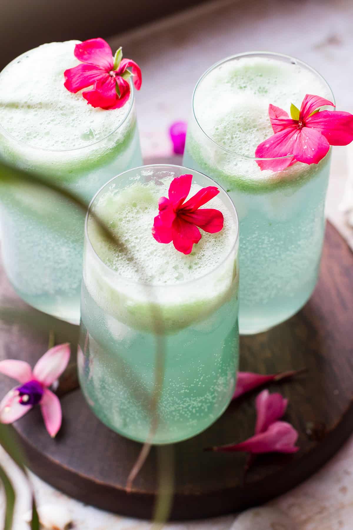 Three tall glasses with turquoise cucumber gin and tonic and fresh pink flowers, some palm leaves blurred out in front.