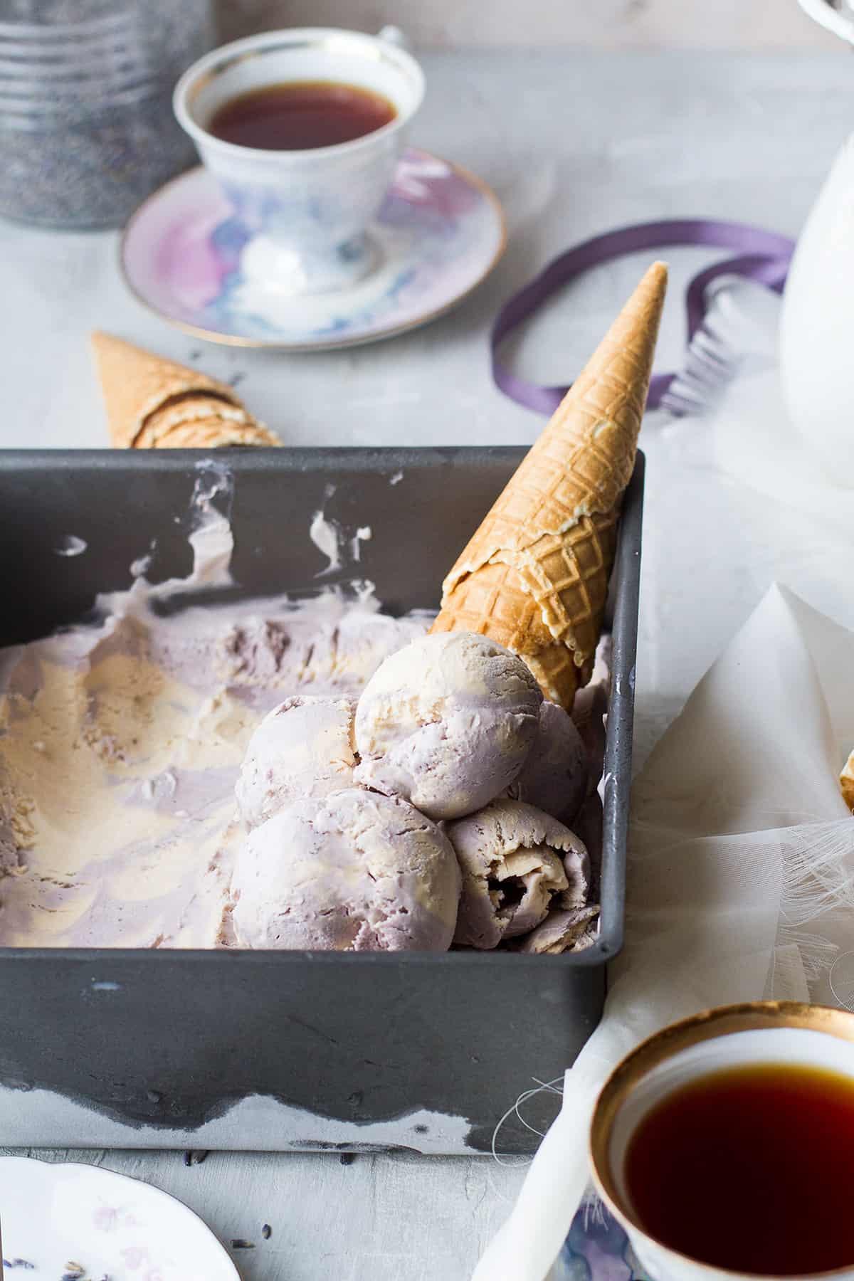 A pan with ice cream in two different colors: purple and beige. Waffle cone in the background.