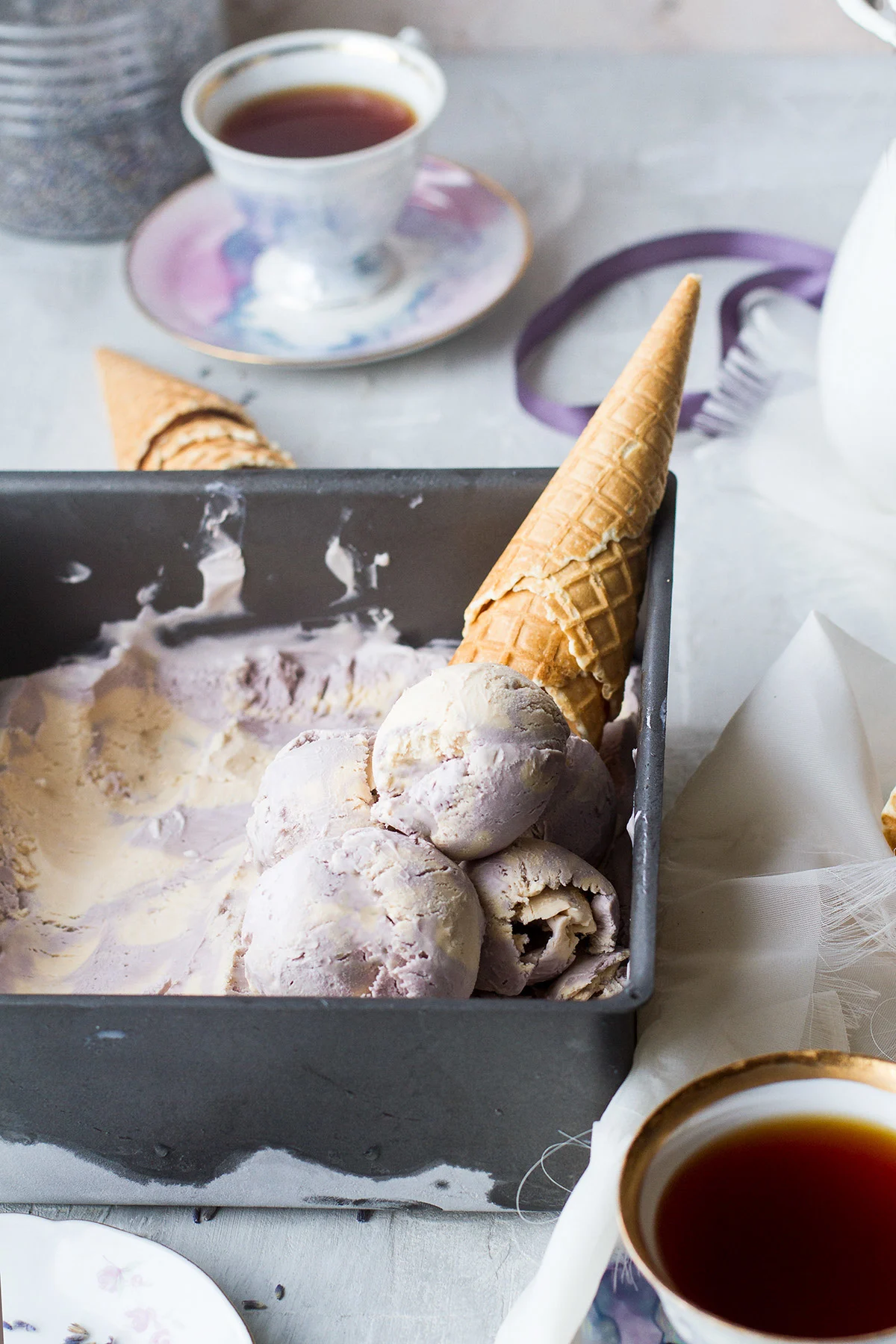 A pan with ice cream in two different colors: purple and beige. Waffle cone in the background.