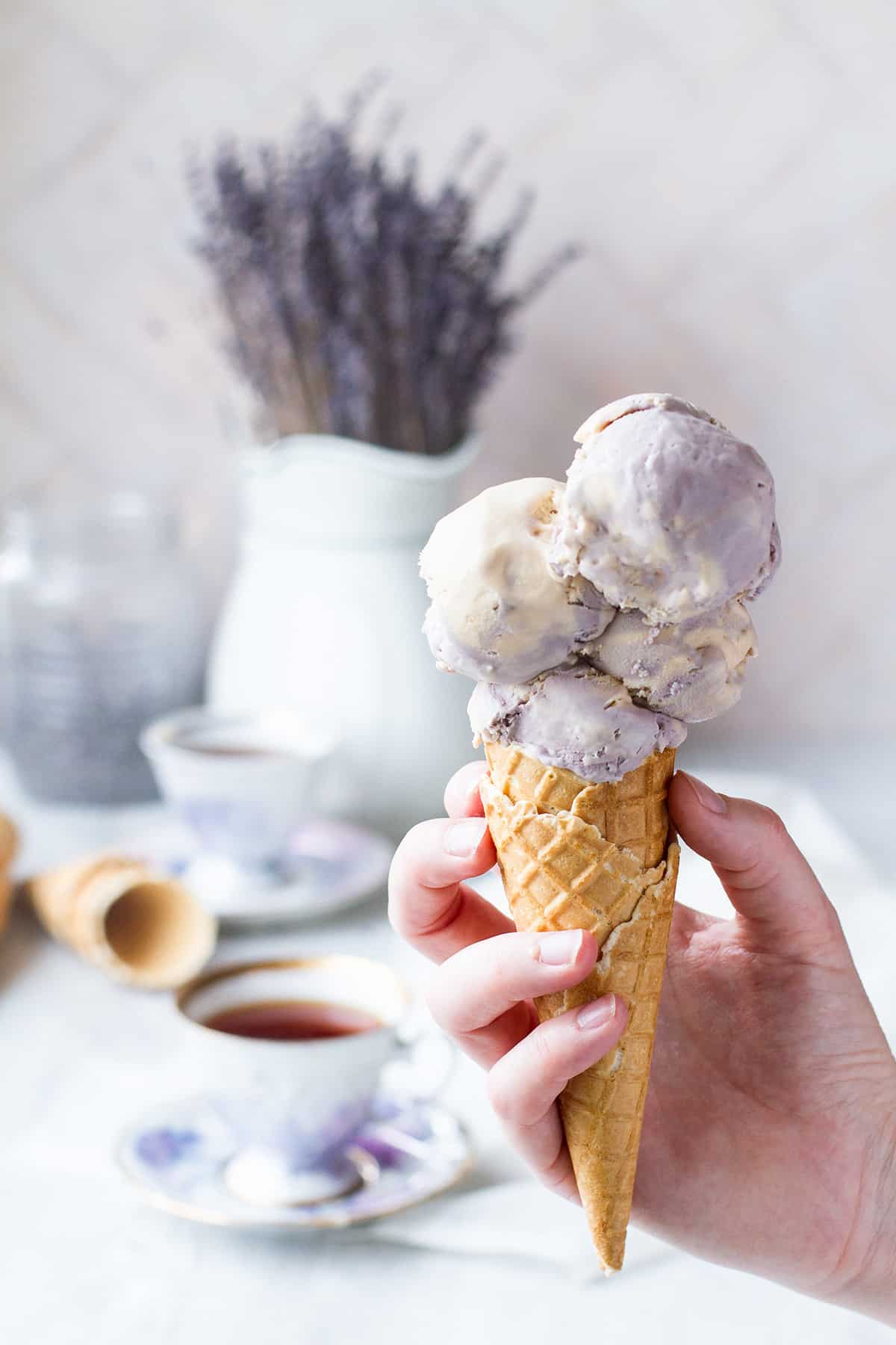 A hand holding a waffle cone with four scoops of ice cream. Earl Grey and lavender flowers in the background.