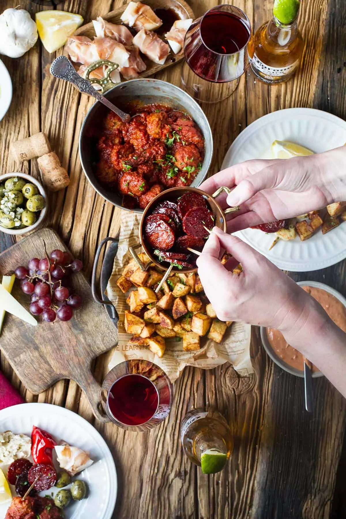 Hand holding red wine chorizo plate over a tapas scene.