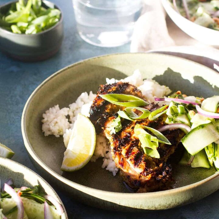 A green shallow dish with grilled chicken thighs over rice and a cucumber salad.