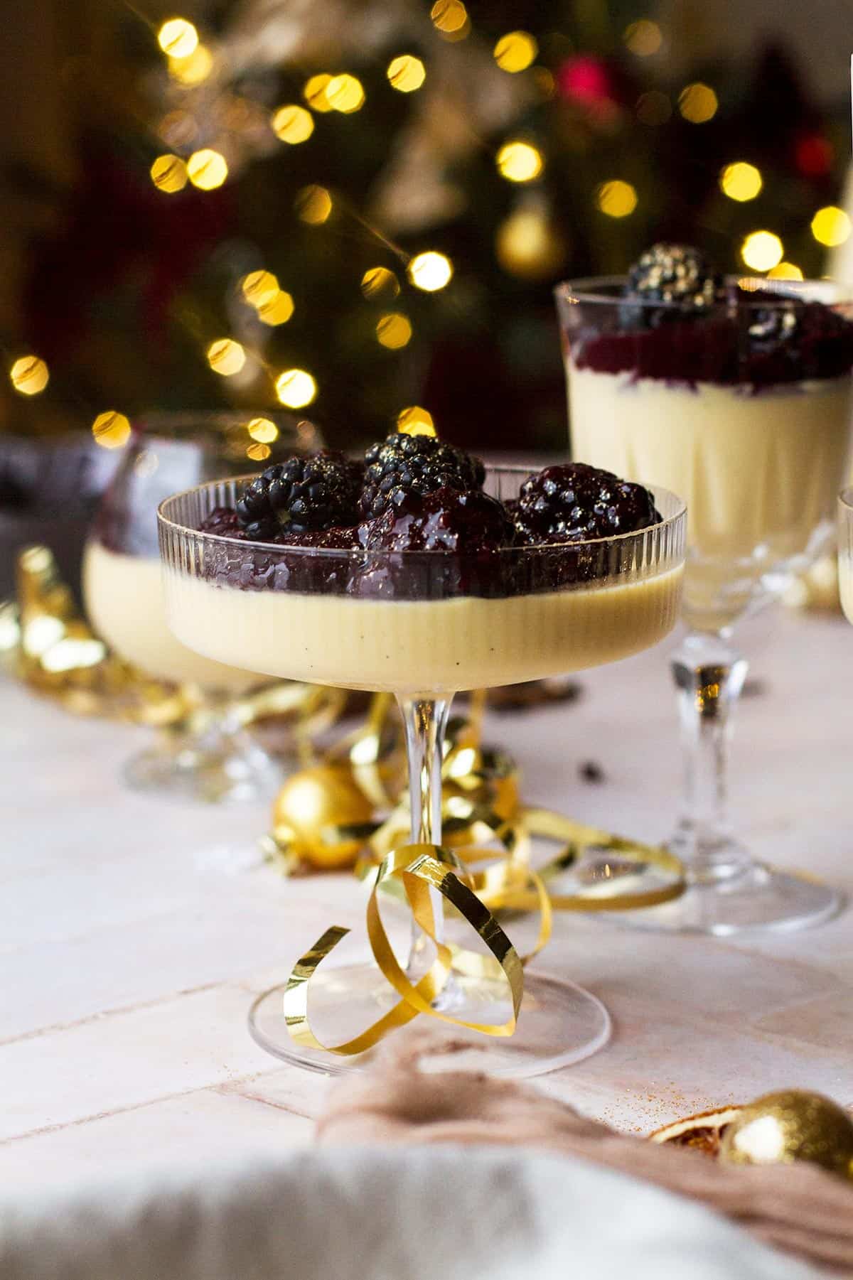 Close-up of a coupe glass with eggnog panna cotta and blackberry sauce, Christmas tree blurred out in the background.