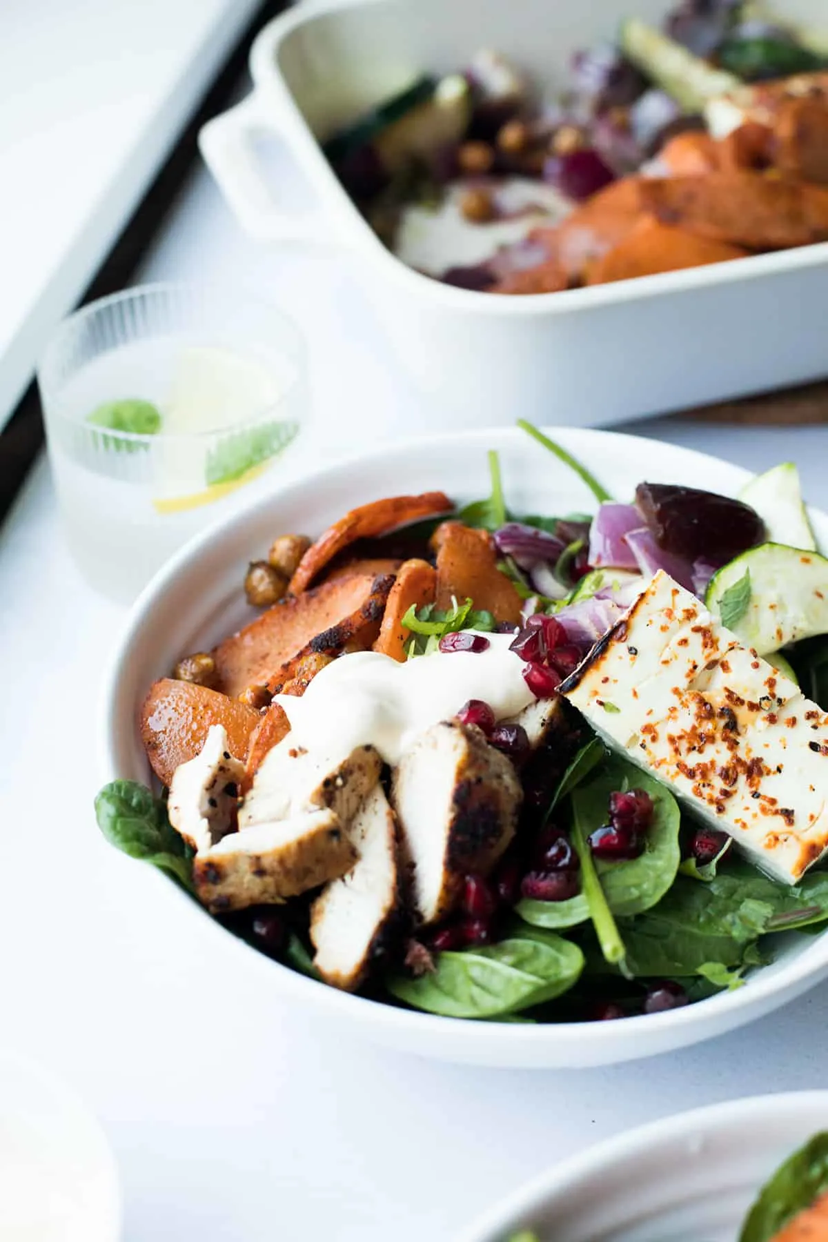 White bowl with salad, chicken, sweet potato and baked feta cheese. Casserole in the background.